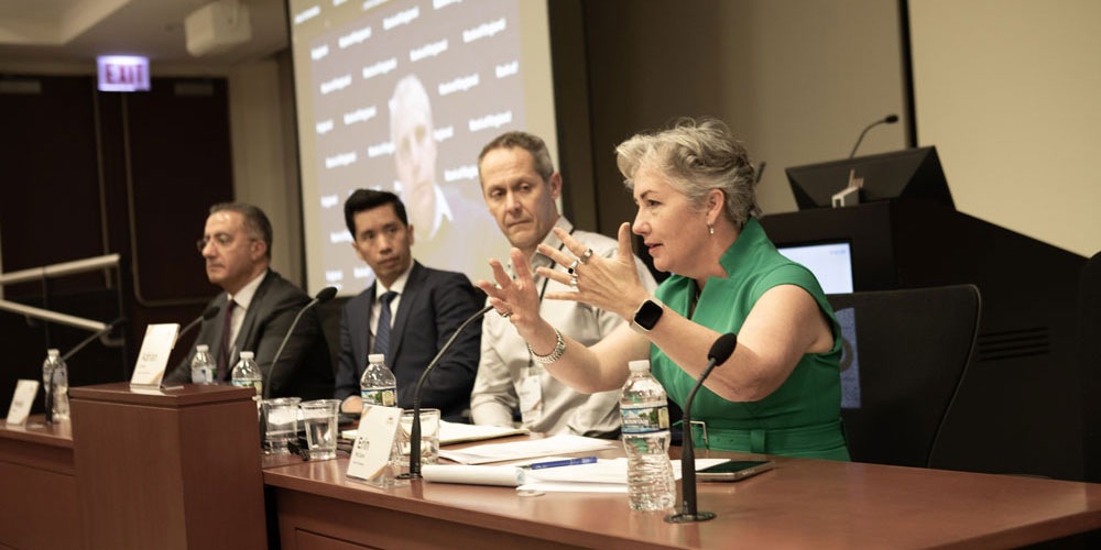 During the 2024 Chicago Payments Symposium, the moderator in a bright green dress introduces a panel session on global payments while three panelists in the room and one participating remotely look on.