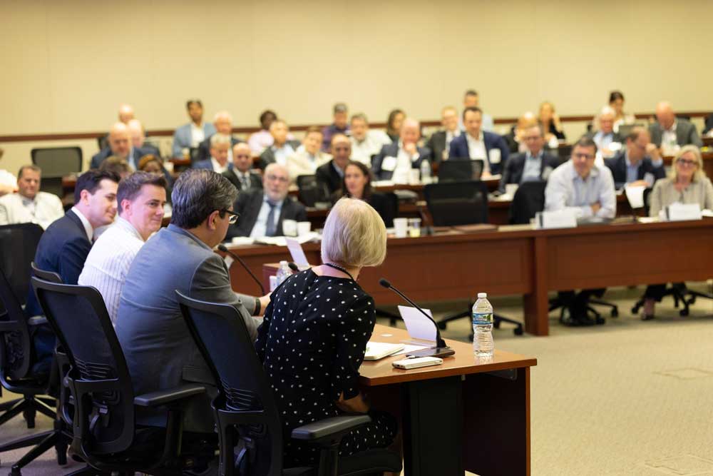 During the 2024 Chicago Payments Symposium, the moderator in a bright green dress introduces a panel session on global payments while three panelists in the room and one participating remotely look on.